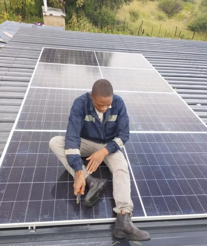 A Pawafactory technician in navy overalls sitting on a row of newly mounted solar panels on a corrugated metal roof